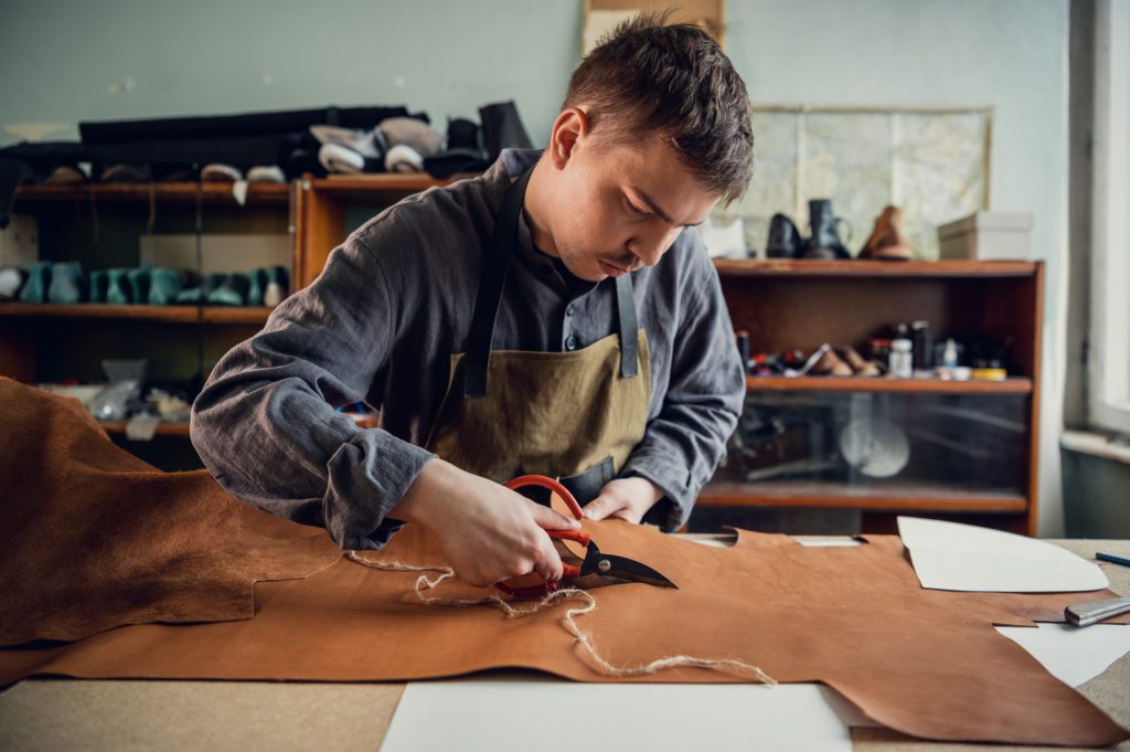 In a boot shop, an experienced young master cuts out leather shoe elements with scissors