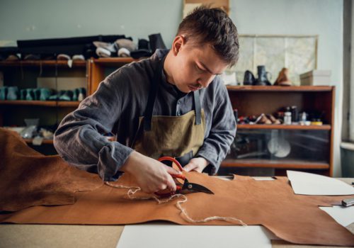 In a boot shop, an experienced young master cuts out leather shoe elements with scissors